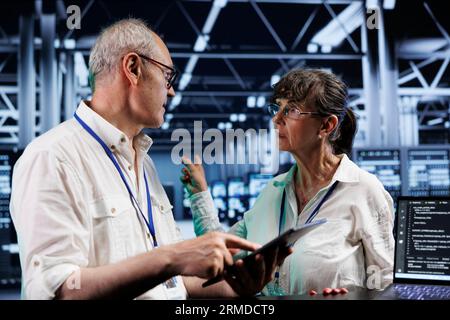 Senior technicians updating server clusters, writing intricate binary code scripts on tablet and laptop. Skilled supervisors using programming to fortify cybersecurity in high tech workplace Stock Photo