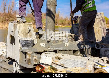 Workers are using shovels to control and directs, fresh concrete into bridge foundation, under construction on river coast. Stock Photo