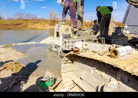 Workers are using shovels to control and directs, fresh concrete into bridge foundation, under construction on river coast. Stock Photo