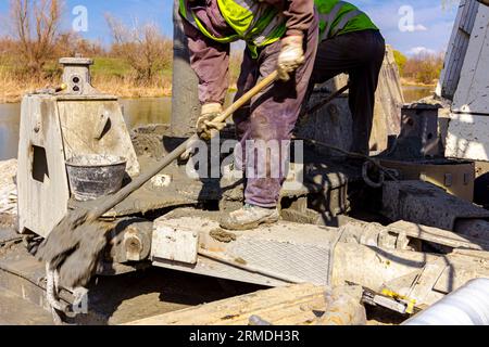 Workers are using shovels to control and directs, fresh concrete into bridge foundation, under construction on river coast. Stock Photo