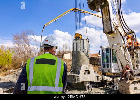 View from behind on construction worker with safety vest and white helmet managing tower drilling machine, big equipment to drill into the ground, for Stock Photo