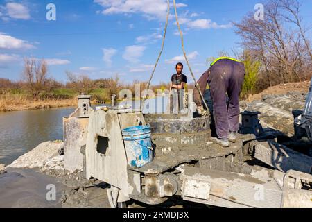 Workers are using shovels to control and directs, fresh concrete into bridge foundation, under construction on river coast. Stock Photo