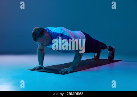 Confident man in sport uniform prosthetic legs doing push-up exercises in horizontal position in neon light on floor. Motivation poster. Stock Photo