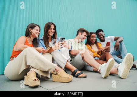 Multiracial smiling teenagers having fun sharing on the social media with their smart phones sitting in row on the floor, blue background . Young multiethnic students enjoying and laughing with funny videos or playing games. Cellphone addiction concept. High quality photo Stock Photo