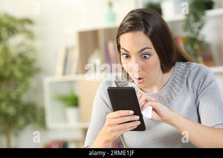 Surprised woman cleaning smart phone with eyes and mouth opened sitting at home in the living room Stock Photo