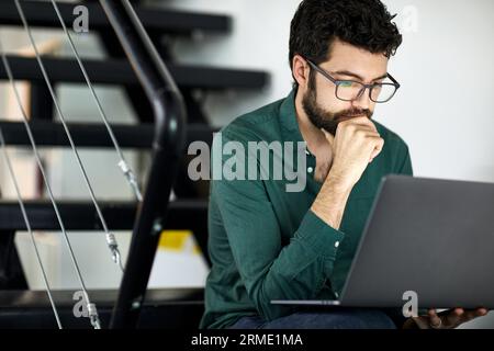 Confident bearded businessman using laptop computer while sitting on steps in office Stock Photo