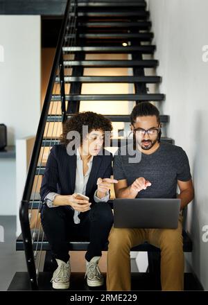 Colleagues discussing over laptop computer while sitting on steps in office Stock Photo