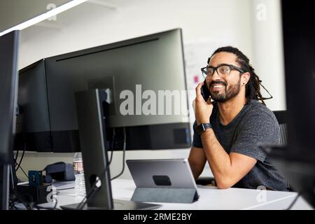 Smiling businessman talking over smart phone while sitting at desk in office Stock Photo