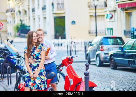 Young romantic couple having a date on a street of Montmartre in Paris, France. Girl is sitting on red scooter and holding traditional French baguette Stock Photo