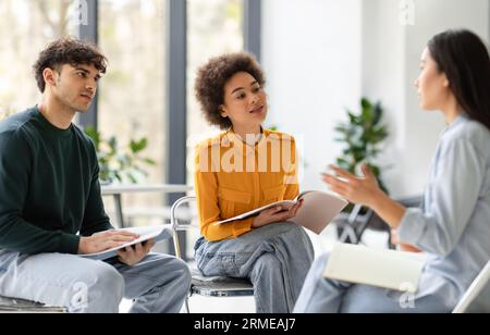 Three international groupmates friends discussing their project, sitting on chairs in circle in university classroom Stock Photo