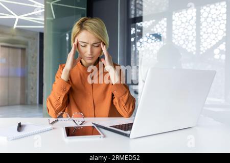 Tired woman at workplace having severe headache, depressed female worker holding hands on head, working inside office with laptop and paper work. Stock Photo