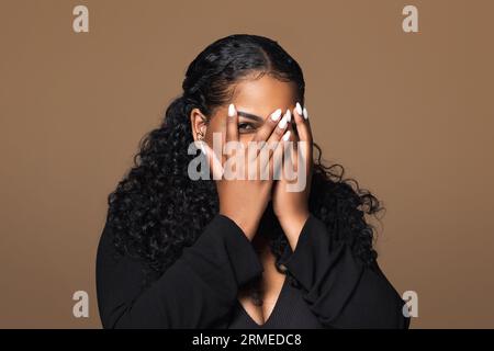Portrait of black chubby woman covering face with hands, feeling afraid or stressed on brown studio background Stock Photo