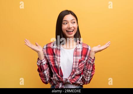 Cheerful japanese lady shrugging shoulders, looking aside and smiling, standing over yellow studio background Stock Photo
