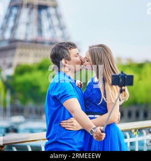 Romantic dating couple on a bridge over the Seine in Paris, taking selfie using selfie stick and kissing, Eiffel tower is in the background Stock Photo