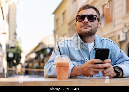 Man wearing denim jacket and sunglasses online messaging using his mobile phone while sitting in open air cafe in summer. Stock Photo