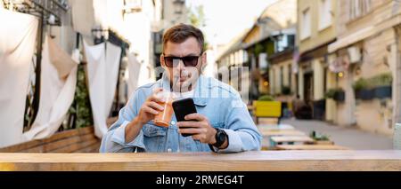 Man wearing denim online messaging using his mobile phone sitting sidewalk cafe and drinking smoothie with straw. Stock Photo