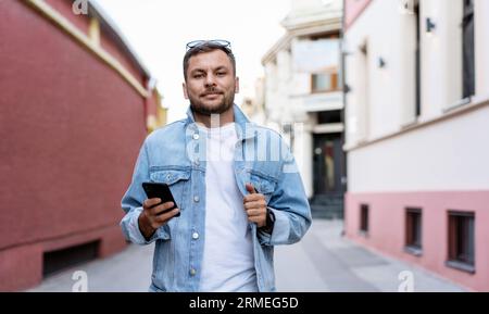Young adult man wearing trendy denim jacket standing on street holding phone in hand and smiling. Stock Photo