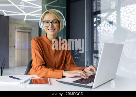 Portrait of successful smiling female programmer, female worker looking at camera using laptop to write new software code, businesswoman in headphones working inside office. Stock Photo
