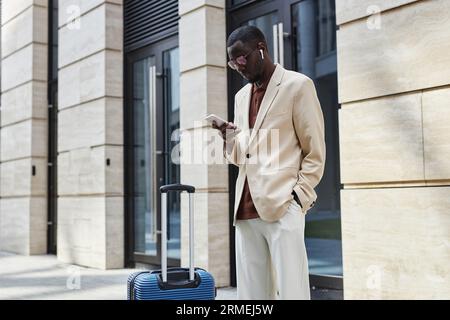 Young serious businessman scrolling or texting in smartphone while standing on sidewalk against modern building and waiting for taxi Stock Photo
