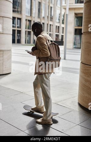 Rear view of young elegant man with backpack and smartphone keeping foot on skateboard while standing in front of camera and texting Stock Photo