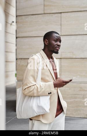Young serene businessman with white textile handbag on shoulder waiting for colleague and using smartphone while standing in front of camera Stock Photo