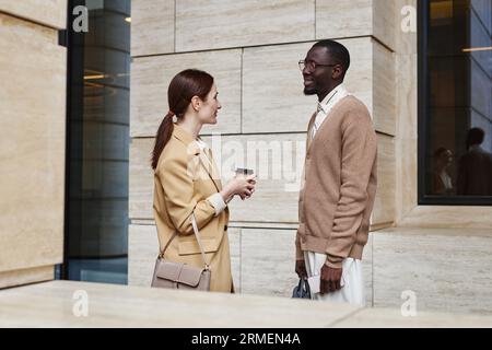 Side view of tow intercultural white collar workers in quiet luxury attire having discussion at coffee break while standing by modern building Stock Photo