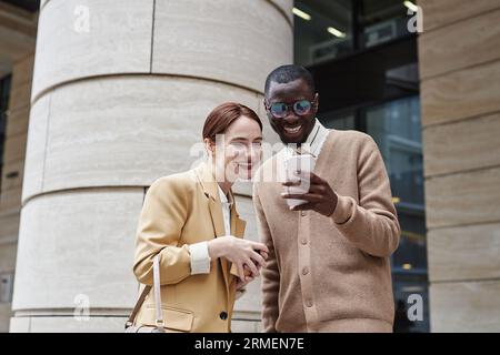 Two young cheerful colleagues in quiet luxury apparel looking at screen of smartphone held by smiling African American male employee Stock Photo