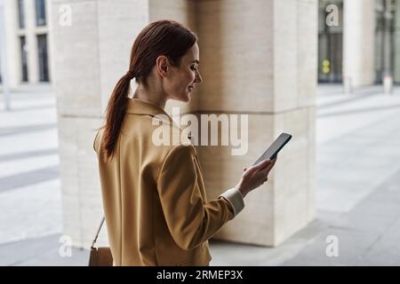 Young brunette female employee looking at smartphone screen while walking in urban environment and communicating in video chat Stock Photo