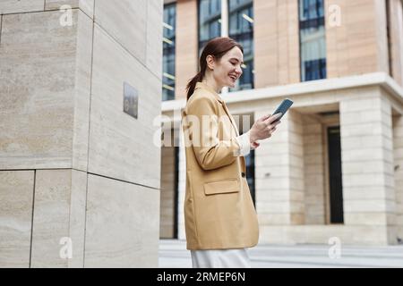 Side view of happy young businesswoman in quiet luxury attire looking at smartphone screen while standing by column of modern architecture Stock Photo