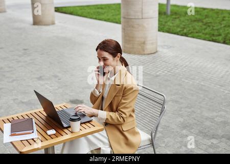 Young smiling female freelancer or solopreneur speaking on mobile phone and looking at laptop screen while sitting by table in urban environment Stock Photo