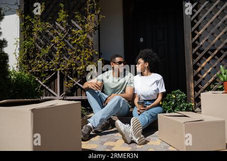 smiling african american man in sunglasses holding coffee and talking to girlfriend near new house Stock Photo