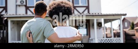 back view of african american couple hugging while standing near new house outdoors, banner Stock Photo