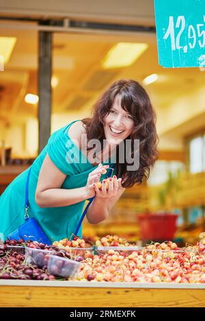 Beautiful French woman choosing ripe organic cherries on a Parisian local market. Customer in food store, supermarket or hypermarket concept Stock Photo