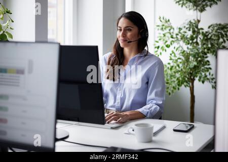Smiling mid adult businesswoman using headset in office in front of computer screen Stock Photo
