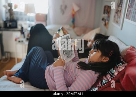 Sisters relaxing in bedroom reading book and using digital tablet Stock Photo