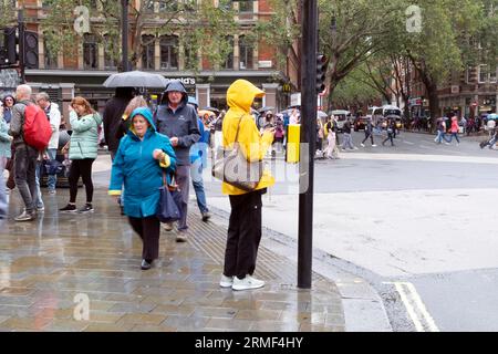 People pedestrians walking in street woman wearing raincoats rain coat on a wet rainy summer day August waiting at traffic lights London KATHY DEWITT Stock Photo
