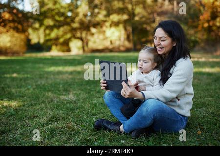 Mother and daughter sitting in autumn park and using digital tablet Stock Photo
