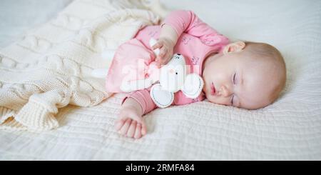 Baby girl having a nap with her favorite mouse toy. Little child sleeping on bed with comforter. Infant kid in sunny nursery Stock Photo