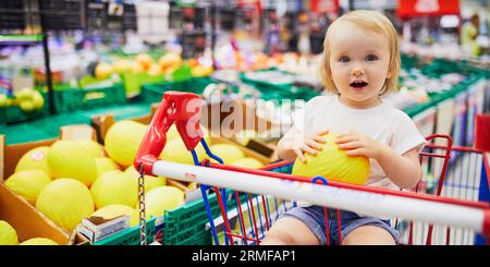 Adorable toddler girl sitting in the shopping cart in a food store or a supermarket. Little kid going shopping Stock Photo