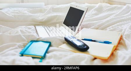 Laptop, notebook and phone in bed on clean white linens. Freelance, distance learning or work from home concept Stock Photo