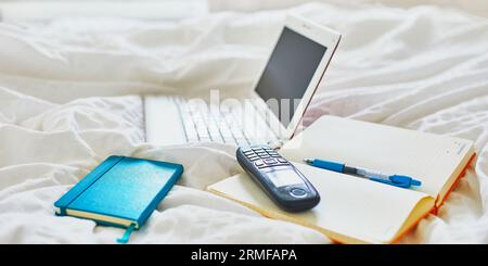 Laptop, notebook and phone in bed on clean white linens. Freelance, distance learning or work from home concept Stock Photo