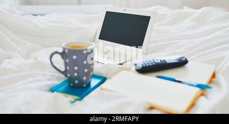 Laptop, cup of coffee, notebook and phone in bed on clean white linens. Freelance, distance learning or work from home concept Stock Photo