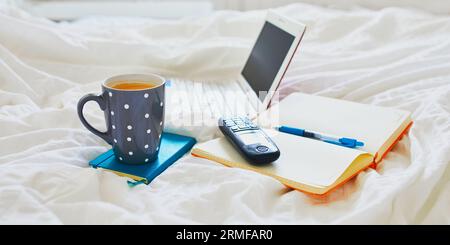 Laptop, cup of coffee, notebook and phone in bed on clean white linens. Freelance, distance learning or work from home concept Stock Photo
