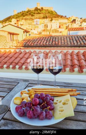Two glasses of red wine with delicious cheese, bread and grapes served on a table of restaurant, cafe or terrace with view to Bosa village, Sardinia, Stock Photo