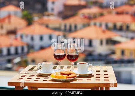 Two glasses of Madeira wine, two cups of fresh espresso coffee and traditional Portuguese sweets in cafe with view to Funchal town, Madeira, Portugal Stock Photo