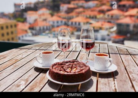 Two glasses of Madeira wine, two cups of fresh espresso coffee and traditional Portuguese honey and nut dessert bolo de mel in cafe with view to Funch Stock Photo