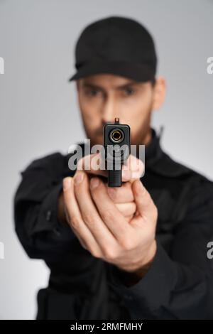 Unrecognizable cop in uniform taking aim, focused on camera indoors. Close up of black weapon, gun, held by blurred police officer silhouette, on gray Stock Photo