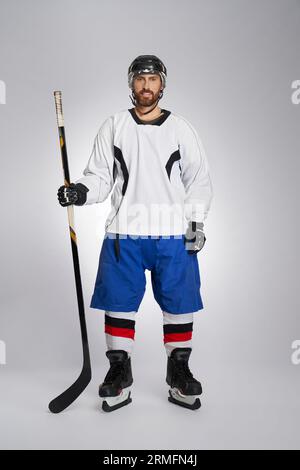 Strong bearded ice hockey player carrying stick and sport bag after game.  Front view of male hockey athlete in full gear looking at camera, isolated  on gray background. Concept of winter team