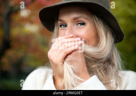 Portrait blonde woman blue eyes in dark hat who laughs and covers her smile with both hands and hair Stock Photo