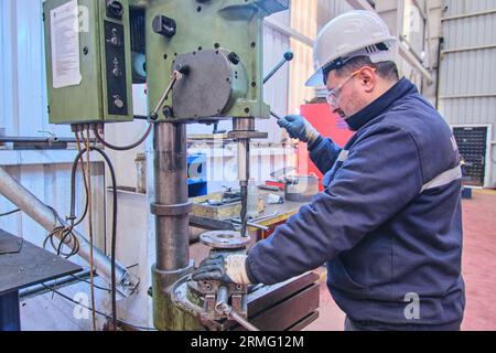 a man in full safety gear like a helmet, visor, and vest. doing welding on a rusted metal pipe. a man using a handheld electrical saw on a metal pipe. Stock Photo